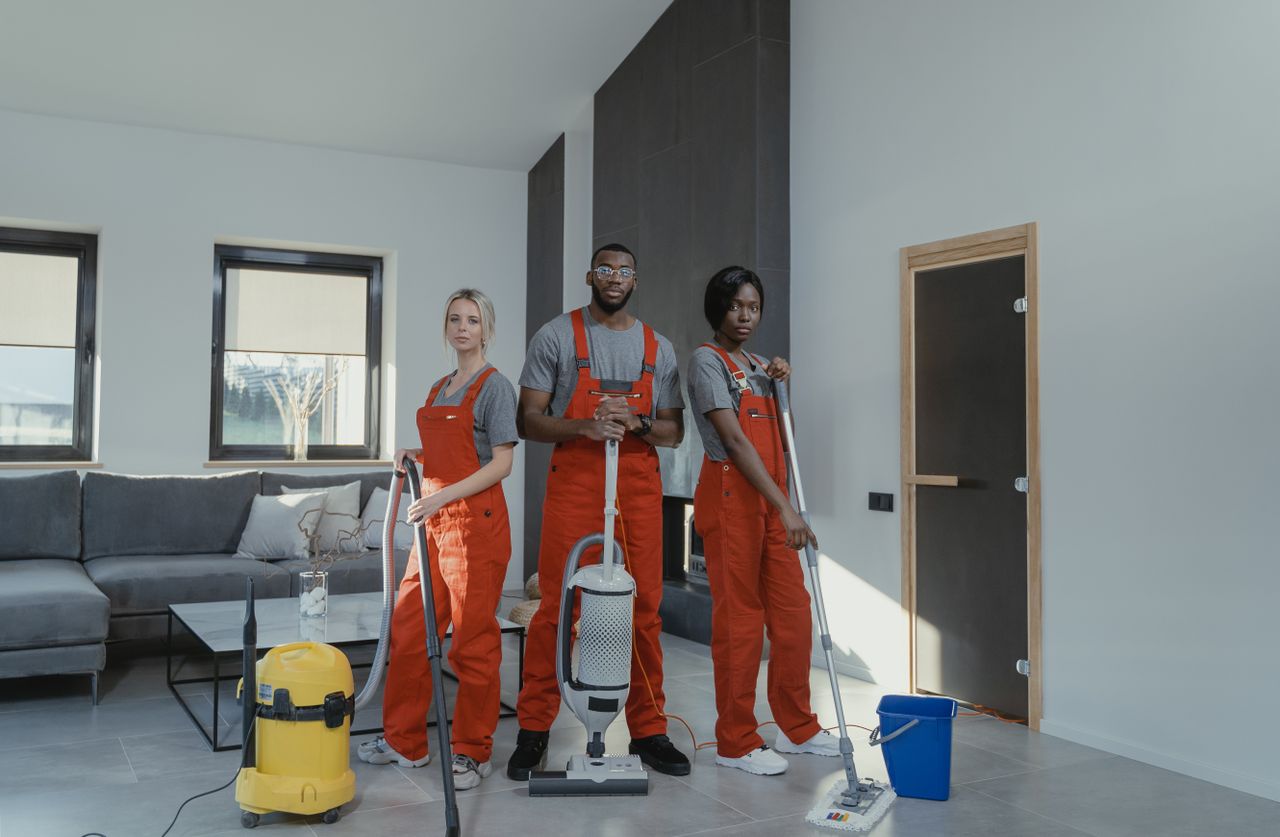 Une équipe de nettoyage diversifiée en uniformes rouges, prête à désinfecter un salon moderne avec aspirateur et serpillière.